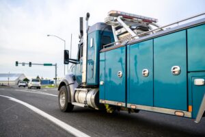 A powerful heavy-duty tow truck with emergency lights and towing equipment drives through a city street, heading to assist a broken-down big rig at a traffic light.